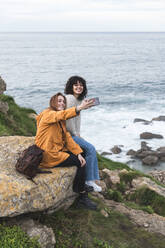 Fröhliche Freundinnen machen ein Selfie, während sie auf einem Felsen am Meer sitzen - JAQF00330
