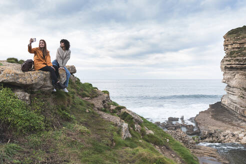 Female friends taking selfie while sitting on cliff by sea at Acantilado El Bolao - JAQF00331