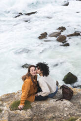 Junge Frau küsst die Wange ihres Freundes, während sie auf einer Klippe am Meer sitzt - JAQF00334