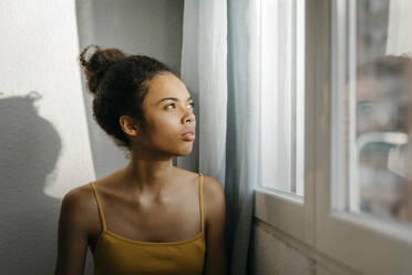 Nachdenkliche junge Frau, die durch das Fenster ihres Hauses schaut - TCEF01613