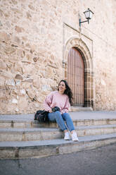 Smiling woman holding camera while sitting on staircase against wall - GRCF00692
