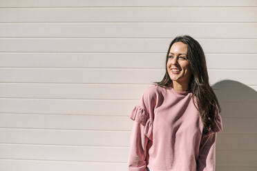 Smiling woman looking away while standing against wall during sunny day - GRCF00696