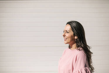 Smiling woman standing by wall during sunny day - GRCF00698