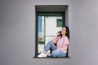 Smiling woman talking on smart phone while sitting on window sill - GRCF00699