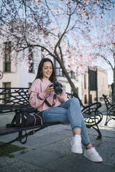 Schöne Frau hält Kamera, während sie auf einer Bank am Fußweg sitzt - GRCF00703