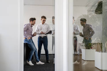 Male entrepreneurs discussing over clipboard seen through doorway at office - DIGF14677