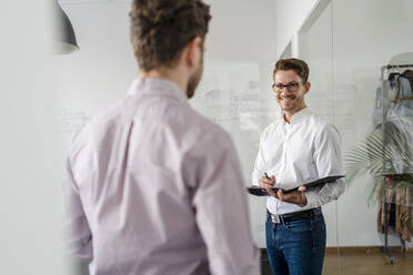 Smiling businessman with clipboard looking at male colleague in office - DIGF14681