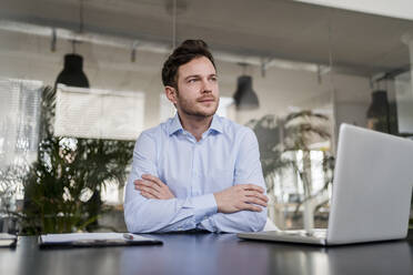 Confident male entrepreneur with arms crossed at desk in office - DIGF14706