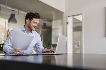 Lächelnder männlicher Unternehmer mit Dokument, der im Büro an einem Laptop arbeitet - DIGF14711