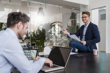 Businessman explaining male colleague working on laptop at desk in office - DIGF14726