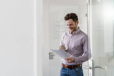 Smiling male entrepreneur with disposable cup analyzing document at office - DIGF14753