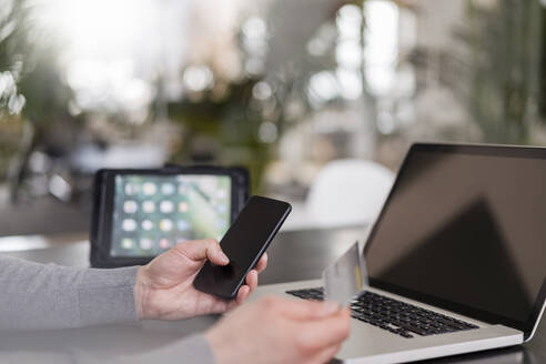 Die Hände eines männlichen Unternehmers beim Online-Bezahlen mit einer Kreditkarte auf einem Smartphone im Büro - DIGF14784