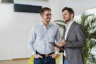 Smiling male colleague with hands in pockets looking at businessman while standing in office - DIGF14796