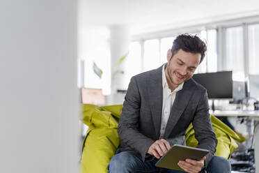 Lächelnder Geschäftsmann, der ein digitales Tablet benutzt, während er auf einem Sitzsack im Kreativbüro sitzt - DIGF14809