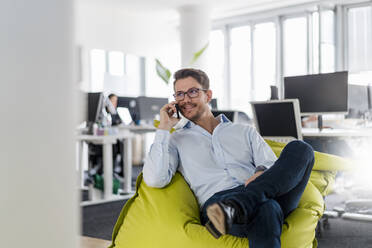 Gutaussehender Geschäftsmann, der über sein Smartphone spricht, während er auf einem Sitzsack im Büro sitzt - DIGF14811