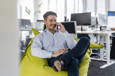 Männlicher Unternehmer, der mit seinem Handy spricht, während er auf einem Sitzsack im Büro sitzt - DIGF14812
