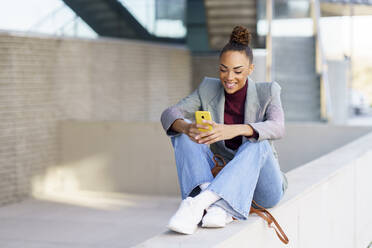 Lächelnde Geschäftsfrau, die ein Smartphone benutzt, während sie auf einer Stützmauer sitzt - JSMF01966