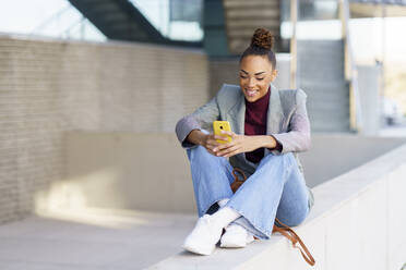 Smiling businesswoman using smart phone while sitting on retaining wall - JSMF01966