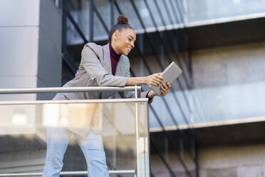 Smiling businesswoman using digital tablet while leaning on railing at office building - JSMF01984