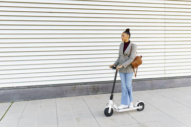 Young woman businesswoman with bag riding electric push scooter against white striped wall - JSMF02000