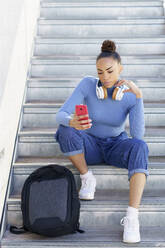 Frau mit Rucksack und Kopfhörern, die ihr Smartphone benutzt, während sie auf einer Treppe sitzt - JSMF02009