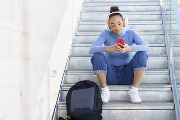 Junge Frau mit Rucksack und Kopfhörern, die auf einer Treppe sitzend ein Mobiltelefon benutzt - JSMF02011