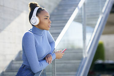 Junge Frau mit Kopfhörern, die wegschaut, während sie am Geländer steht - JSMF02012