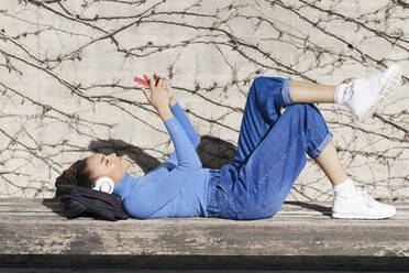 Young woman with headphones using mobile phone while resting on bench during sunny day - JSMF02028