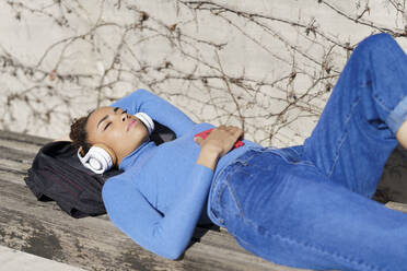 Young woman wearing headphones napping while lying on bench during sunny - JSMF02029