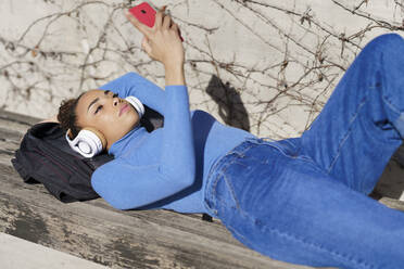 Young woman with headphones headphones using mobile phone while resting on bench - JSMF02030