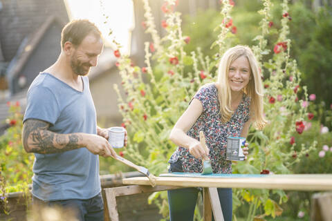 Lächelnde blondhaarige Frau, die mit einem im Garten stehenden Mann ein Holzbrett bemalt, lizenzfreies Stockfoto