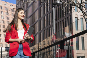 Young businesswoman in red blazer walking with laptop in city - PNAF00881