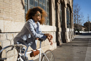Afro woman in sunglasses leaning on bicycle against building - RCPF00741