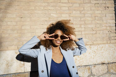 Smiling young woman in sunglasses gesturing while standing against wall - RCPF00750