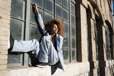 Smiling young woman balancing while sitting on window sill - RCPF00753