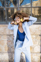 Young woman enjoying music through headphones while standing against building - RCPF00757