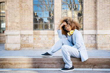 Confident woman with headphones sitting on footpath against building - RCPF00759