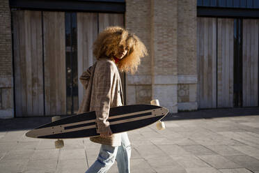 Afro-Frau, die ein Skateboard trägt, während sie an einem sonnigen Tag auf dem Fußweg gegen ein Gebäude läuft - RCPF00762