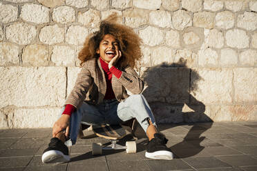 Lachende Afro-Frau, die auf einem Skateboard an der Wand sitzt, an einem sonnigen Tag - RCPF00777