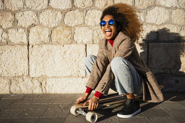 Fröhliche Frau mit Sonnenbrille, die auf einem Skateboard an der Wand sitzt, an einem sonnigen Tag - RCPF00778