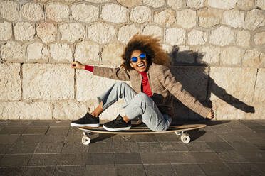 Fröhliche Frau mit ausgestreckten Armen, die auf einem Skateboard an der Wand sitzt, an einem sonnigen Tag - RCPF00779