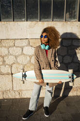 Coole Einstellung Frau trägt Sonnenbrille mit Skateboard stehen gegen Gebäude während des sonnigen Tages - RCPF00781
