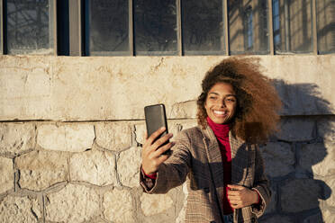 Lächelnde junge Frau, die ein Selfie mit ihrem Smartphone macht, während sie an einem Gebäude steht - RCPF00789