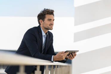 Geschäftsmann mit digitalem Tablet schaut weg, während er sich auf ein Geländer auf einer Büroterrasse stützt - SBOF02844