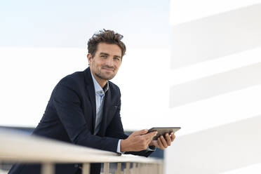 Lächelnder Geschäftsmann, der ein digitales Tablet hält, während er an einem Geländer auf einer Büroterrasse steht - SBOF02845