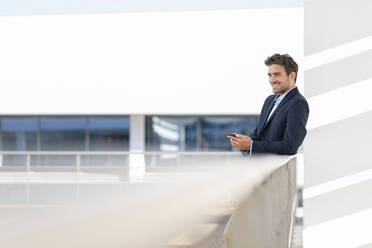 Smiling businessman with mobile phone looking away while standing by railing at office terrace - SBOF02848