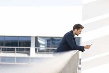 Businessman using smart phone while leaning on railing at office terrace - SBOF02849