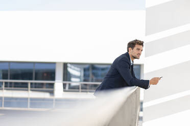 Mittlerer erwachsener Geschäftsmann mit Mobiltelefon, der wegschaut, während er sich auf ein Geländer auf einer Büroterrasse stützt - SBOF02850