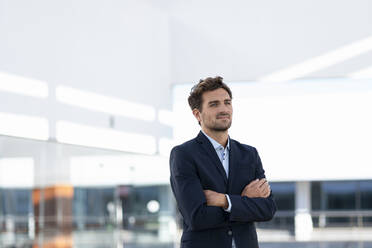 Thoughtful businessman looking away while standing with arms crossed against office building - SBOF02851