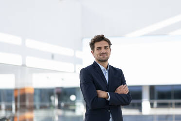 Businessman smiling while standing with arms crossed against office building - SBOF02852